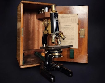 May include: A vintage black microscope with brass accents, in a wooden case with a green felt lining. The microscope has two lenses and a stage for viewing specimens.
