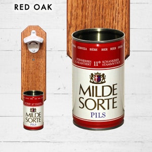 May include: A wooden wall mounted bottle opener with a red oak finish. The bottle opener has a white metal bottle opener attached to it. There is a beer can holder below the bottle opener. The beer can holder is holding a red and white beer can with the words "Milde Sorte Pils" printed on it.