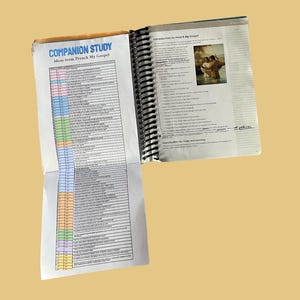 Puede incluir: Libro abierto con espiral titulado "Companion Study Ideas from Preach My Gospel". La página izquierda tiene una lista codificada por colores. La página derecha tiene texto y una imagen de dos personas en el agua. El fondo es amarillo claro liso.