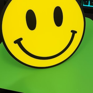May include: A large, round yellow smiley face with black eyes and a curved black mouth. The smiley face is set against a bright green background.