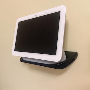 Puede incluir: Una pantalla inteligente blanca con una pantalla negra está montada en una pared. La pantalla está sujeta por un estante negro y curvado. La pared está pintada de color beige claro.