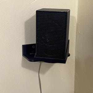 Op de afbeelding: Een zwarte rechthoekige luidspreker staat op een zwarte muurbeugel. De luidspreker heeft een mesh-achtige textuur. Een grijze kabel loopt van de luidspreker. De muur is beige.