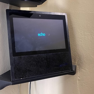 Puede incluir: Un dispositivo doméstico inteligente negro con pantalla, montado en un estante negro contra una pared beige. La pantalla muestra la palabra "echo" en turquesa. El dispositivo tiene una rejilla de altavoz en la parte inferior.