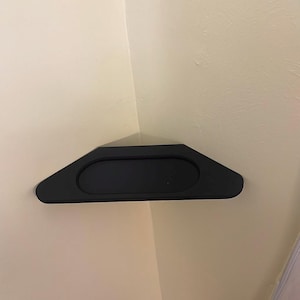 Puede incluir: Una estantería de esquina negra con un centro rectangular redondeado. La estantería está montada en una pared de color beige claro. La estantería está diseñada para encajar en una esquina, maximizando el espacio.