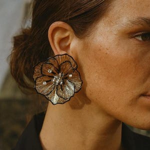 May include: A large, gold-toned flower-shaped earring with black outlining and small, sparkling embellishments. The earring is worn on the ear, showcasing its intricate design and metallic finish. The jewelry piece is a statement accessory.