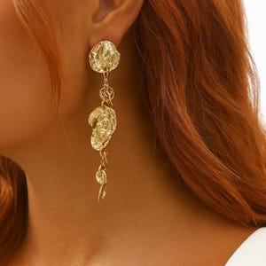 Peut inclure: Boucles d'oreilles pendantes dorées avec un design texturé et organique. Les boucles d'oreilles présentent un gros clou irrégulier, relié par des chaînes à trois éléments texturés supplémentaires. Les boucles d'oreilles sont présentées sur un fond de cheveux brun-rouge.