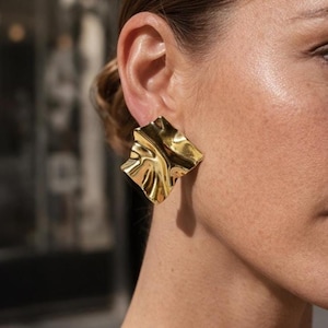 May include: A close-up of a gold-colored, square-shaped earring with a rippled, textured surface. The earring is worn on the ear of a person with fair skin and dark hair. The background is blurred.