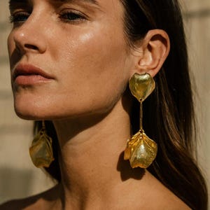 Puede incluir: Pendientes dorados con forma de pétalo. Los pendientes tienen un tallo largo y delgado que conecta dos elementos florales distintos. El diseño presenta una estética texturizada y orgánica, con un acabado pulido. Los pendientes son una pieza llamativa, adecuada para ocasiones especiales.