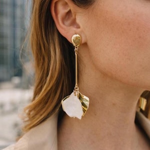 May include: Gold-toned drop earrings featuring a petal design. The earrings have a post backing with a long, slender gold bar that connects to a white and gold petal-shaped charm. The earrings are worn on a person.