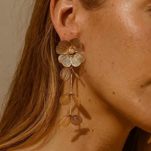 May include: Gold-toned floral drop earrings. The earrings feature a large flower at the top with three smaller, delicate flower-shaped pendants hanging below. The earrings have a textured, detailed design and are worn on the ear.