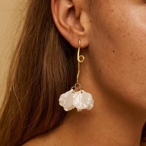 Puede incluir: Un pendiente dorado con un diseño de alambre largo y curvado. El pendiente presenta un grupo de colgantes blancos en forma de pétalo en la parte inferior. El pendiente se lleva en la oreja de una persona.