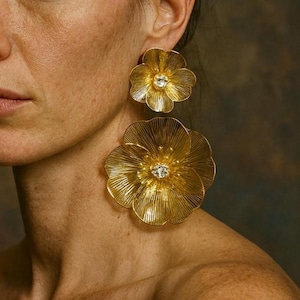 Puede incluir: Pendientes dorados con forma de flor y una piedra central brillante. Los pendientes son grandes y detallados, con un diseño texturizado y acanalado. Una pieza llamativa.