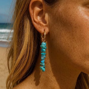 Puede incluir: Pendientes de aro dorados con una fila vertical de piedras turquesas. Los pendientes cuelgan, con las piedras turquesas de varios tamaños y formas. El fondo muestra una playa y el mar.