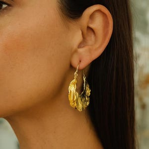 May include: Close-up of a gold-colored earring with a textured, leaf-like design. The earring is a hoop style, with a hook closure. The jewelry is worn on the ear of a person with dark hair.