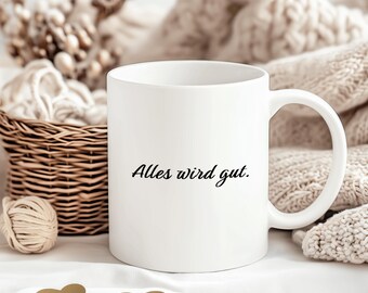 May include: White ceramic mug with a black inscription that reads "Alles wird gut." The mug is sitting on a white surface with gold heart-shaped decorations.