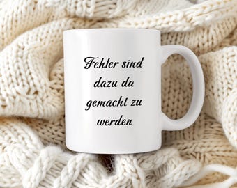 Puede incluir: Taza de cerámica blanca con texto negro que dice "Fehler sind dazu da gemacht zu werden". La taza está sobre una manta de punto blanca.