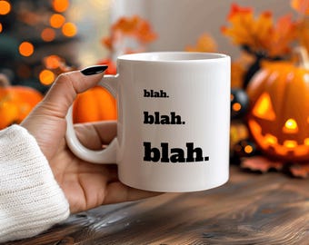 May include: White ceramic mug with black text that reads "blah. blah. blah." The mug is being held by a hand in a white sweater. The background is blurry and features a pumpkin and autumn foliage.