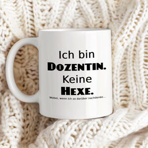 Könnte beinhalten: Weiße Keramik-Tasse mit schwarzem Text: "Ich bin DOZENTIN. Keine Hexe. Wobei, wenn ich so darüber nachdenke...". Die Tasse hat einen weißen Henkel und steht auf einer cremefarbenen Strickdecke.
