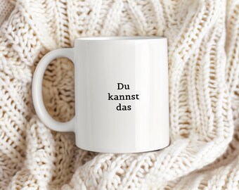 May include: White ceramic coffee mug with a black inscription that reads "Du kannst das" on a white knitted background.