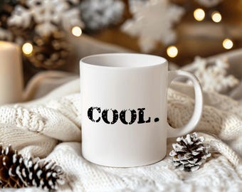 May include: White ceramic mug with black text that reads "COOL." The mug is sitting on a white knitted blanket with pine cones.