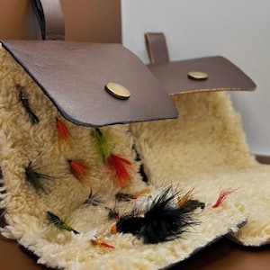 May include: Two brown leather fly fishing wallets with brass snap closures. The interior of each wallet is lined with thick, cream-coloured sheepskin wool, which holds an assortment of colourful feathered fishing flies in various sizes and styles.
