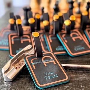 Puede incluir: Etiquetas de equipaje negras con bordes naranjas y turquesas, con el texto "Vina Tram" y el logotipo "Fore! Amigos". Cada etiqueta tiene una correa de cuero negro con una remache dorado. Una herramienta de palo de golf plateada está en primer plano.