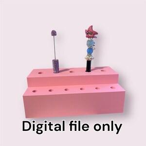May include: A pink, two-tiered display stand with multiple holes for holding items. Two decorative items are in the stand: a purple, bedazzled pen and a pen with a starfish topper. The text "Digital file only" is at the bottom.