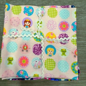 Puede incluir: Un conjunto de cuatro cuadrados de tela rosa con un borde blanco festoneado. Cada cuadrado presenta un patrón colorido diferente con círculos, estrellas, flores y personajes de dibujos animados. Los patrones incluyen una princesa, un carruaje, un castillo y una rana.