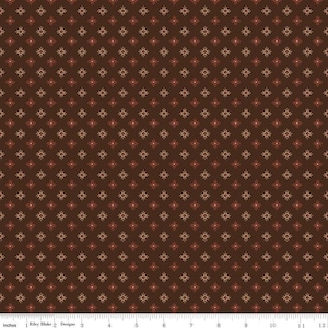 May include: Brown fabric with a repeating pattern of small, stylized sunbursts in shades of red and tan. The fabric has a dark brown background and is marked with inches along the bottom edge. The text "Riley Blake Designs" is also visible.