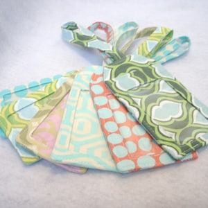 May include: A collection of fabric luggage tags in various patterns and colors. The tags feature designs with teal, green, pink, and orange accents. Each tag has a loop for attachment. The patterns include polka dots and geometric shapes.