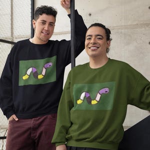 Puede incluir: Dos personas con sudaderas con un gráfico de un gusano con rayas moradas, amarillas, negras y blancas sobre un fondo verde. Una sudadera es negra y la otra es verde.