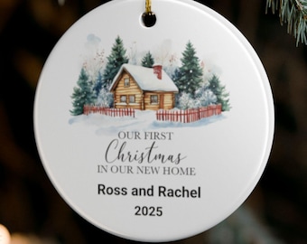 May include: A white ceramic Christmas bauble featuring a watercolour scene of a cabin in a snowy setting. The text reads "Our First Christmas in Our New Home", with the names "Ross and Rachel" and the year "2025".