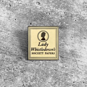 May include: A gold-colored metal pin with a black silhouette of a woman's head and the text "Lady Whistledown's Society Papers" on a cream background.