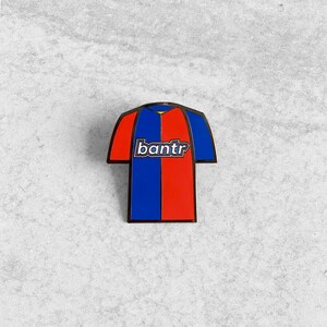 May include: A blue and red enamel pin shaped like a soccer jersey with the word "bantr" printed on the front.