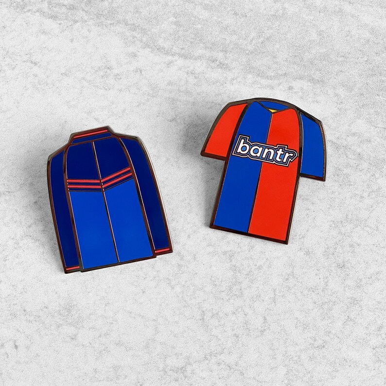 May include: Two enamel pins, one blue and one red and blue. The blue pin is a jacket with a red stripe down the front. The red and blue pin is a soccer jersey with the word "bantr" on the front.