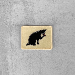 Könnte beinhalten: Ein schwarzer Kater, der auf einem beigefarbenen Stoffpatch gestickt ist. Die Katze sitzt und schaut nach rechts. Der Patch hat einen weißen Rand.