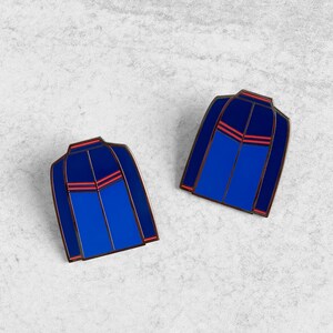 May include: Two blue enamel pins shaped like a jacket with red stripes. The pins are on a light gray background.