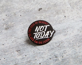 May include: Black enamel pin with white text that reads "What do we say to the god of death? Not today." The pin has a red outline.
