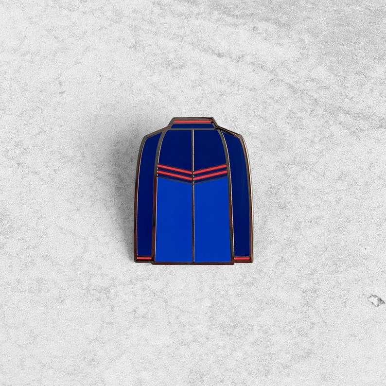 May include: Blue enamel pin with red accents depicting a jacket with a zipper and a V-shaped design on the front.