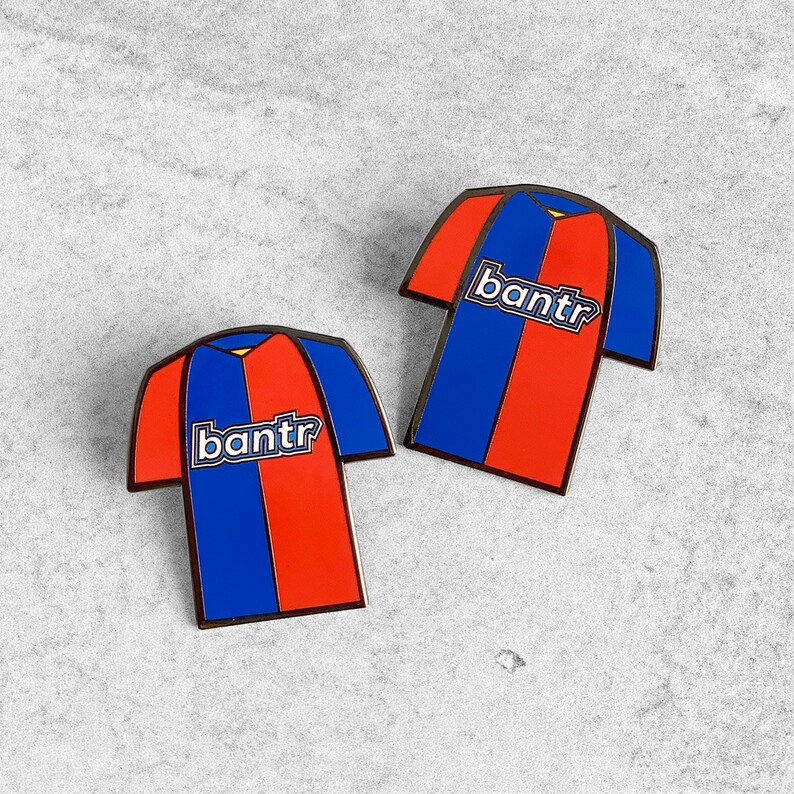May include: Two enamel pins shaped like soccer jerseys with a red and blue color scheme. The jerseys have the word "bantr" printed on the front.