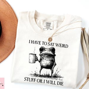 Peut inclure: T-shirt crème avec le texte "I HAVE TO SAY WEIRD STUFF OR I WILL DIE." Une illustration en noir et blanc d'une grenouille tenant une tasse est centrée sous le texte. Un collier doré est drapé sur le t-shirt.