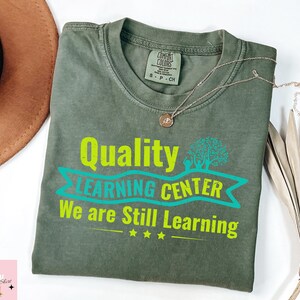Könnte beinhalten: Ein olivgrünes T-Shirt mit dem Text "Quality Learning Center We are Still Learning" in leuchtendem Grün und Türkis. Das Shirt hat einen Rundhalsausschnitt und kurze Ärmel. Eine goldene Halskette liegt über dem Shirt.