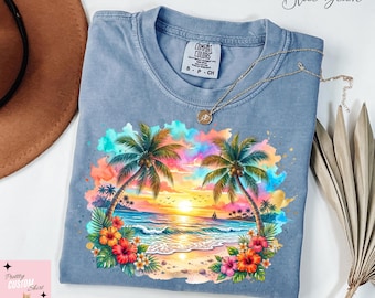 Puede incluir: Camiseta azul vaquero con una escena de playa tropical. El gráfico presenta palmeras, una puesta de sol, olas del océano y flores coloridas. Un collar de oro descansa sobre la camisa. La camisa tiene una etiqueta Comfort Colors.