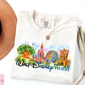 K&ouml;nnte beinhalten: Cremefarbenes T-Shirt mit einem farbenfrohen Aquarelldesign von Walt Disney World-Wahrzeichen. Das Design umfasst ein Schloss, Spaceship Earth und andere Attraktionen, mit den Worten "Walt Disney World" in schwarzer und blauer Schrift.