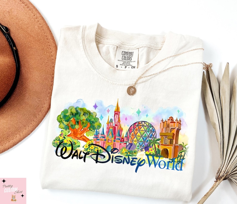 K&ouml;nnte beinhalten: Cremefarbenes T-Shirt mit einem farbenfrohen Aquarelldesign von Walt Disney World-Wahrzeichen. Das Design umfasst ein Schloss, Spaceship Earth und andere Attraktionen, mit den Worten "Walt Disney World" in schwarzer und blauer Schrift.