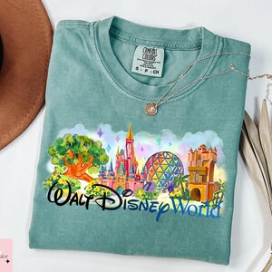 K&ouml;nnte beinhalten: Ein teal Comfort Colors T-Shirt mit einer farbenfrohen Grafik von Walt Disney World-Wahrzeichen, darunter ein Schloss, Spaceship Earth und der Tree of Life. Die Worte "Walt Disney World" sind in einer stilisierten Schriftart gedruckt.