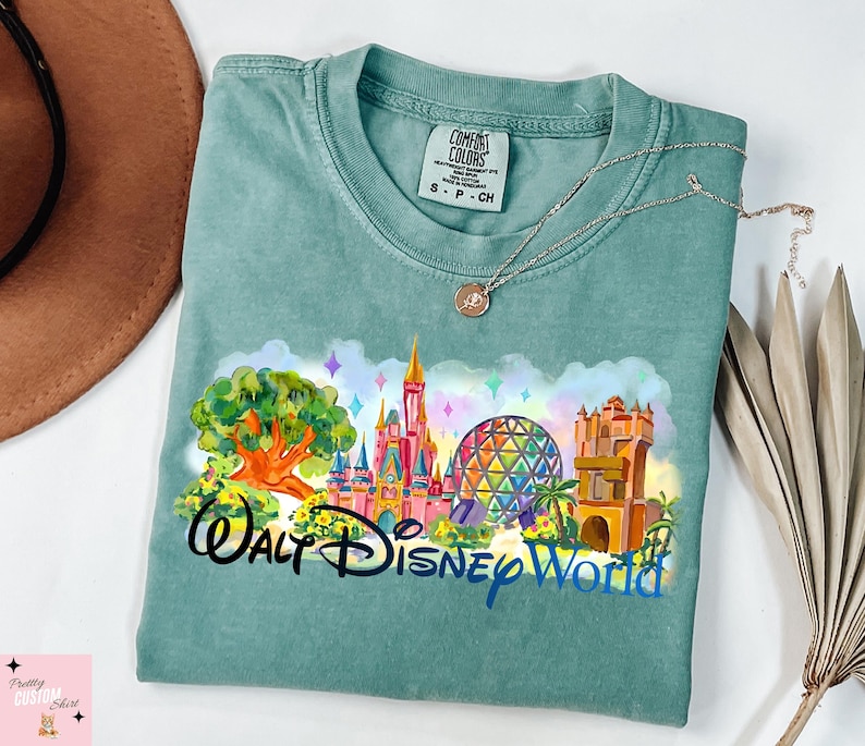 K&ouml;nnte beinhalten: Ein teal Comfort Colors T-Shirt mit einer farbenfrohen Grafik von Walt Disney World-Wahrzeichen, darunter ein Schloss, Spaceship Earth und der Tree of Life. Die Worte "Walt Disney World" sind in einer stilisierten Schriftart gedruckt.
