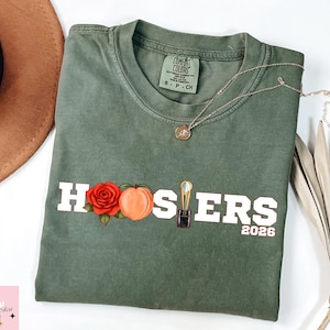 May include: Olive green t-shirt with the word "HOOSIERS" in white block letters, featuring a red rose, peach, and trophy graphic. The year "2026" is printed below. A gold necklace rests on the shirt.