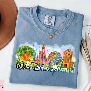 K&ouml;nnte beinhalten: Hellblaues T-Shirt mit einem farbenfrohen Grafikdesign von Walt Disney World-Wahrzeichen, darunter ein Schloss, Spaceship Earth und der Tower of Terror. Die Worte "Walt Disney World" sind darunter gedruckt. Eine goldene Halskette liegt auf dem Shirt.