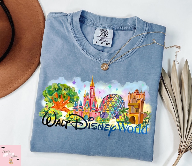 K&ouml;nnte beinhalten: Hellblaues T-Shirt mit einem farbenfrohen Grafikdesign von Walt Disney World-Wahrzeichen, darunter ein Schloss, Spaceship Earth und der Tower of Terror. Die Worte "Walt Disney World" sind darunter gedruckt. Eine goldene Halskette liegt auf dem Shirt.